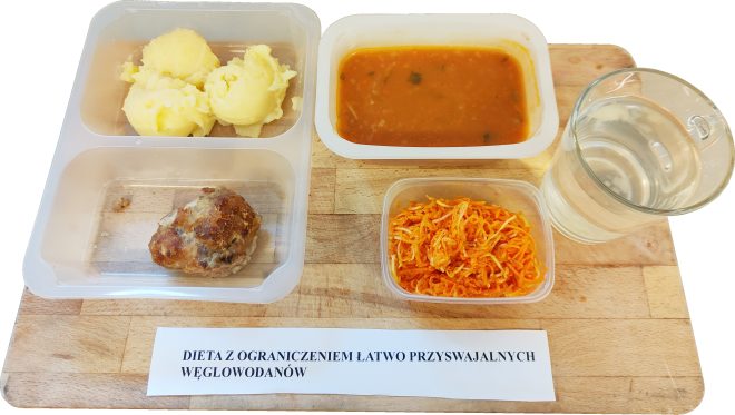 Na drewnianej tacy znajduje się szpitalny posiłek opisany jako „Dieta z ograniczeniem łatwo przyswajalnych węglowodanów”. W pojemnikach ułożono porcję puree ziemniaczanego, kotlet mielony, zupę pomidorową z ryżem, surówkę z marchwi i selera oraz szklankę wody. Całość stanowi przykład obiadu dietetycznego podawanego w placówce medycznej