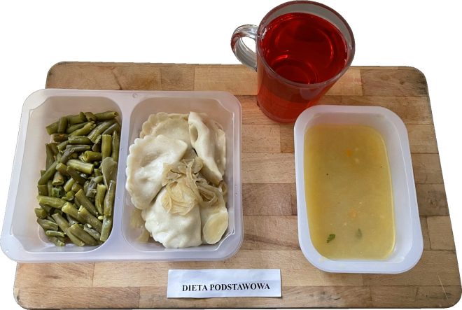 Zestaw obiadowy w pojemnikach dietetycznych, podpisany jako dieta podstawowa. W skład posiłku wchodzą pierogi z podsmażoną cebulką, fasolka szparagowa oraz zupa jarzynowa. Do picia czerwony napój w szklance. Całość ustawiona na drewnianej tacy