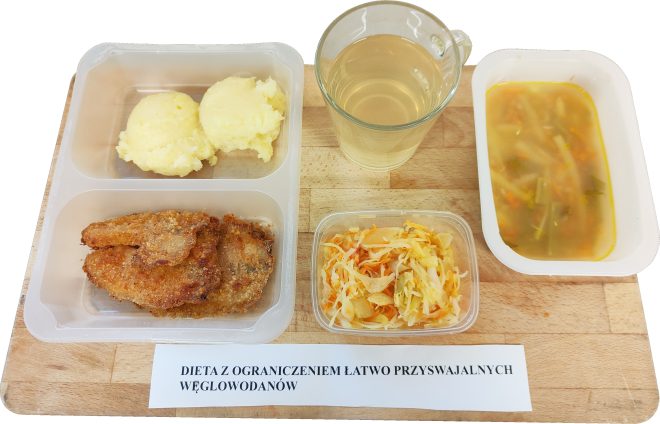 Na tacy widać zestaw obiadowy dla pacjenta przebywającego na diecie z ograniczeniem łatwo przyswajalnych węglowodanów. W skład posiłku wchodzi zupa jarzynowa z drobnymi kawałkami warzyw, filet drobiowy w panierce, ziemniaki puree oraz surówka z białej kapusty i marchewki. Do picia podano herbatkę ziołową lub owocową. Posiłek jest zbilansowany, lekkostrawny i dostosowany do potrzeb osób wymagających ograniczenia węglowodanów prostych