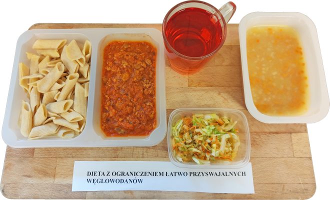 Na tacy znajduje się zestaw obiadowy w diecie z ograniczeniem łatwo przyswajalnych węglowodanów. Posiłek składa się z makaronu rurki, sosu pomidorowego z mięsem, surówki z kapusty i marchwi oraz kubka z czerwonym napojem owocowym. W oddzielnym pojemniku podano lekką zupę jarzynową. Na tacy widnieje etykieta „Dieta z ograniczeniem łatwo przyswajalnych węglowodanów”