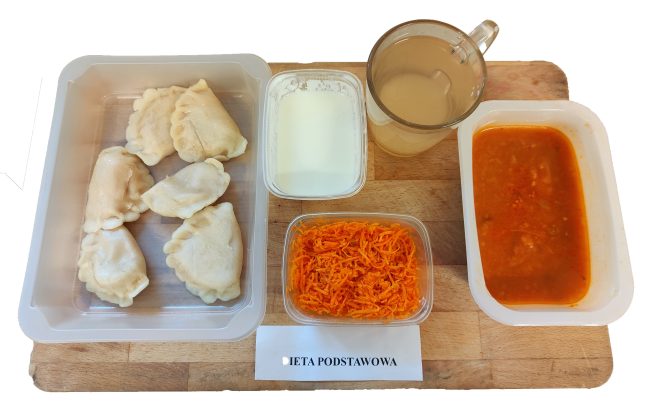 Zestaw obiadowy w diecie podstawowej. Na tacy widać pięć pierogów w pojemniku, obok biały kubek z kompotem oraz pojemniki z dodatkami: biały sos lub śmietana, surówka z marchewki i zupa pomidorowa z ryżem. Wszystko ułożone na drewnianej desce, z karteczką z napisem „Dieta podstawowa”