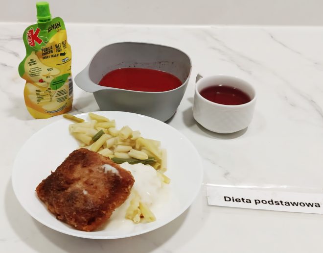 Na białym talerzu znajduje się panierowany kotlet rybny podany z żółtą fasolką szparagową i sosem. Obok miseczka z czerwonym barszczem, kubek z kompotem oraz saszetka z musem owocowym Kubuś. Przy zestawie umieszczona jest etykieta z napisem „Dieta podstawowa”