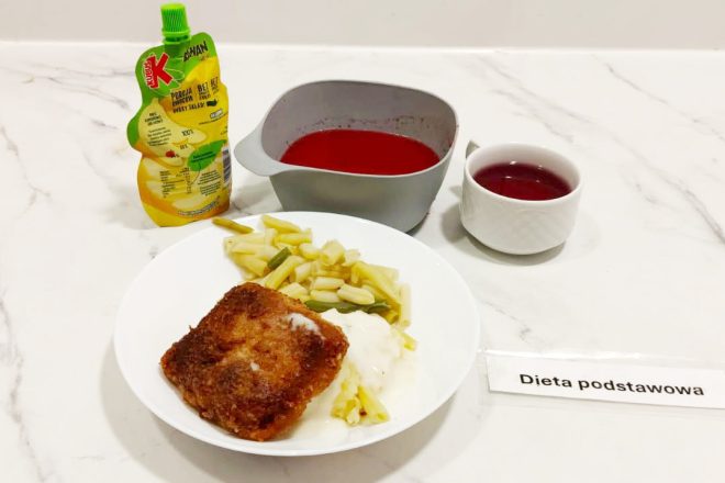 Obiad w diecie podstawowej: panierowany kotlet z ziemniakami puree i gotowaną fasolką szparagową, podany na białym talerzu. Obok miseczka z czerwonym barszczem, kubek z napojem owocowym oraz saszetka musu Kubuś. Całość podana na białym blacie z karteczką „Dieta podstawowa”