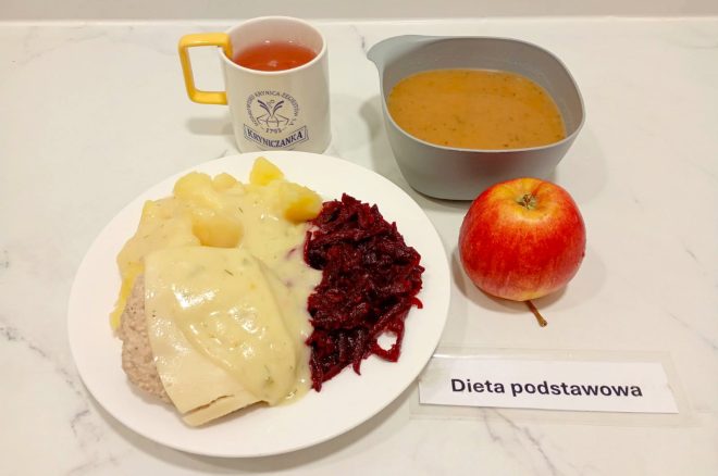 Na zdjęciu widoczny jest zestaw obiadowy oznaczony jako „Dieta podstawowa”. Na białym talerzu znajduje się porcja ziemniaków polanych sosem koperkowym, gotowany mielony kotlet przykryty plasterkiem sosu oraz porcja buraczków na ciepło. Obok ustawiono szarą miskę z zupą o pomarańczowym kolorze, czerwoną jabłko oraz kubek z herbatą w białym naczyniu z żółtym uchem i logo „Kryniczanka”. Całość ustawiona jest na jasnym blacie