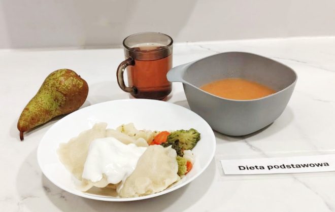 Zestaw obiadowy w diecie podstawowej. Na talerzu pierogi polane śmietaną z dodatkiem gotowanych warzyw: brokułów, kalafiora i marchewki. Obok szara miska z zupą pomidorową, szklanka herbaty oraz gruszka. Na białej kartce napis „Dieta podstawowa”