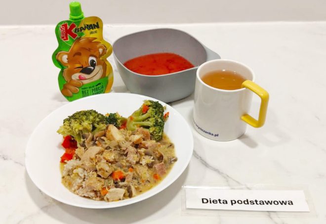 Zestaw obiadowy oznaczony jako „Dieta podstawowa”. Na białym talerzu porcja kaszy z duszonym mięsem, pieczarkami, marchewką i brokułami. Obok w miseczce czerwona zupa, w kubku herbata, a z lewej strony mus owocowy w opakowaniu z wizerunkiem misia