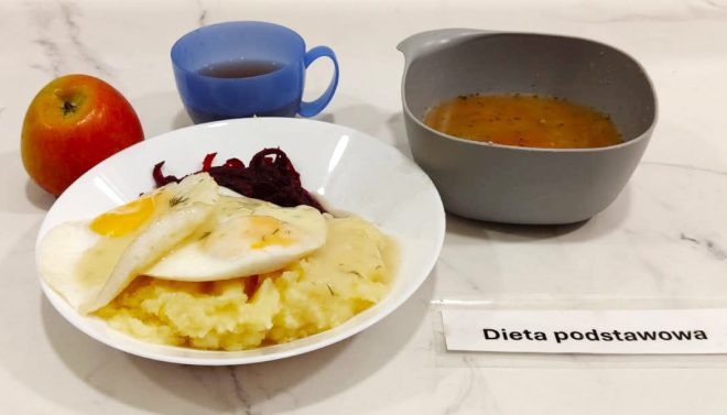 Na białym talerzu porcja puree ziemniaczanego z dwoma jajkami sadzonymi i sosem koperkowym, obok buraczki. Po lewej stronie czerwone jabłko i kubek z ciemnym napojem, po prawej miska z zupą jarzynową. Całość ustawiona na jasnym blacie z karteczką „Dieta podstawowa”