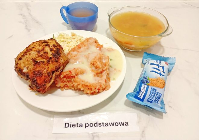 Na tacy znajduje się zupa jarzynowa w miseczce, napój w kubku, porcja ryżu z sosem, panierowane udko z kurczaka oraz surówka z kapusty. Obok położono batonik zbożowy o smaku jagodowym. Kartka informuje: „Dieta podstawowa”