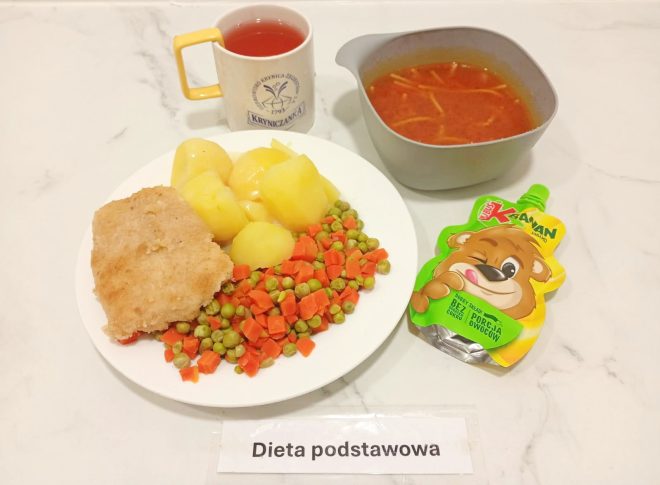 Zestaw obiadowy obejmuje zupę pomidorową z makaronem, gotowane ziemniaki, gotowaną marchewkę z groszkiem, panierowany filet z ryby oraz napój w kubku. Dodatkowo na tacy znajduje się owocowy mus w saszetce. Na kartce przed talerzem widnieje napis „Dieta podstawowa”