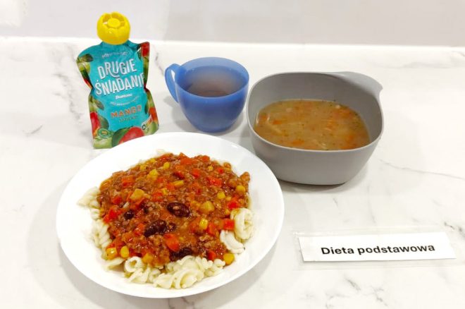 Zestaw obiadowy w ramach diety podstawowej: na białym talerzu podano makaron świderki z sosem mięsno-warzywnym z kukurydzą, czerwoną papryką i fasolą. Do posiłku dołączono zupę jarzynową w szarej miseczce, szklankę z herbatą oraz owocowy mus w saszetce o smaku mango i jabłka. Obok widnieje karteczka z napisem „Dieta podstawowa”