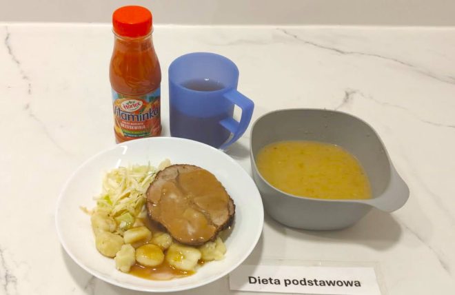 Na talerzu porcja pieczeni z sosem, kopytka oraz surówka z kapusty. Obok szara miseczka z zupą jarzynową, kubek z napojem i butelka soku marchwiowo-brzoskwiniowego. Po prawej stronie kartka z napisem „Dieta podstawowa”