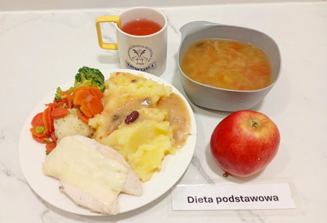 Na zdjęciu znajduje się zestaw obiadowy w ramach diety podstawowej. Na białym talerzu podano gotowaną pierś drobiową z serem, puree ziemniaczane z sosem pieczarkowym oraz gotowane warzywa – marchewkę, brokuły i kalafior. Obok stoi miseczka z zupą jarzynową, czerwone jabłko oraz kubek z herbatą owocową w żółto-białym kubku z logo „Kryniczanka”. Całość oznaczona napisem „Dieta podstawowa”
