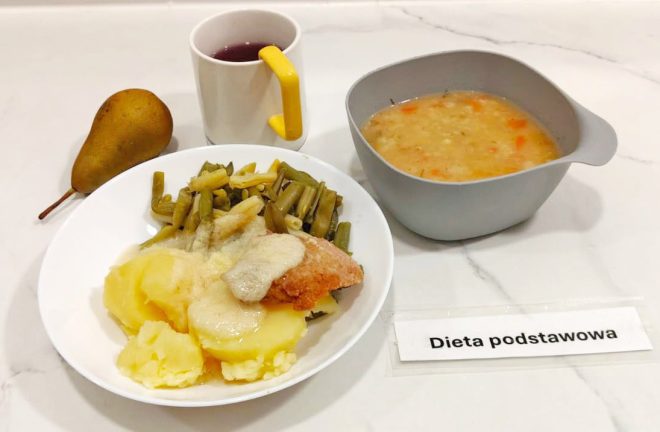 Na białym talerzu porcja ziemniaków, zielona fasolka szparagowa i kawałek ryby polany sosem. Obok szara miseczka z zupą jarzynową i kubek z czerwonym kompotem. Z lewej strony leży gruszka. Na stole kartka z napisem „Dieta podstawowa”
