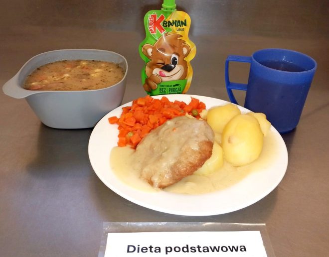 Na talerzu trzy ziemniaki, gotowana marchewka z groszkiem i kotlet w sosie. Obok szara miska z zupą oraz saszetka musu owocowego Kubuś o smaku jabłko-banan. Do picia – napój w niebieskim kubku. Na stole leży karteczka z napisem „Dieta podstawowa”