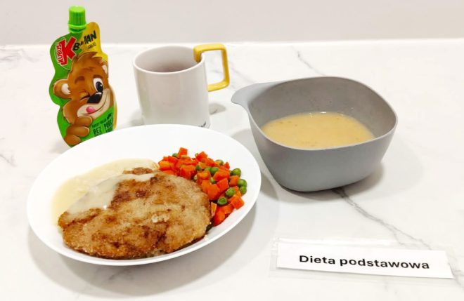 Na talerzu znajduje się panierowany kotlet schabowy z sosem, podany z gotowaną marchewką z groszkiem. Obok widać szarą miseczkę z zupą oraz kubek z napojem. Dodatkiem jest mus owocowy w tubce. Całość stanowi posiłek w diecie podstawowej