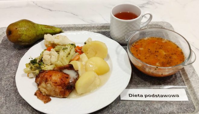 Na tacy z napisem „Dieta podstawowa” znajduje się talerz z gotowanymi ziemniakami, pieczonym udkiem kurczaka w sosie oraz gotowanymi warzywami – brokułem, kalafiorem i marchewką. Obok stoją miska z zupą jarzynową o czerwonym kolorze, filiżanka z herbatą oraz gruszka