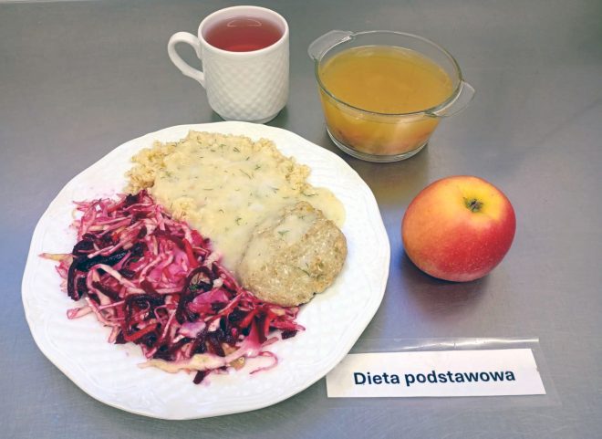 Na tacy z napisem „Dieta podstawowa” znajduje się talerz z kotletem mielonym w sosie koperkowym, kaszą oraz surówką z czerwonej kapusty i buraczków. Obok ustawiono miseczkę z zupą jarzynową o żółtym kolorze, kubek z herbatą i jabłko