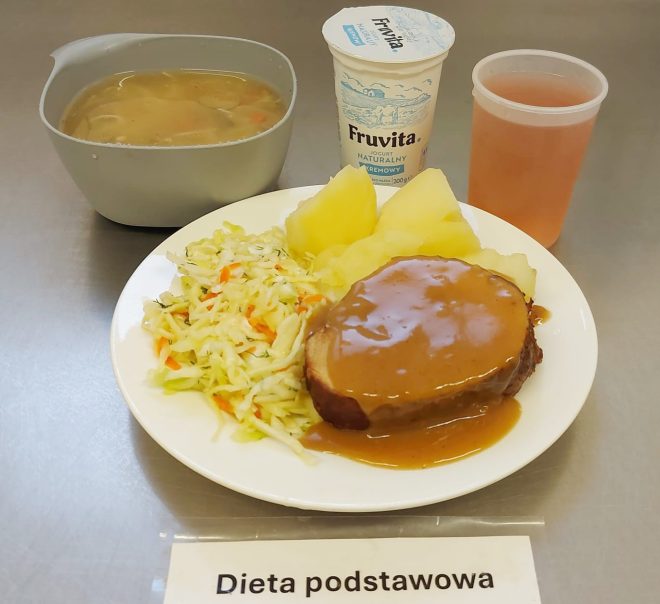 Obiad składający się z kawałka pieczeni wieprzowej polanej sosem, gotowanych ziemniaków i surówki z kapusty. Obok zupa jarzynowa w misce, kubek z kompotem i jogurt naturalny. Na stoliku kartka z napisem Dieta podstawowa