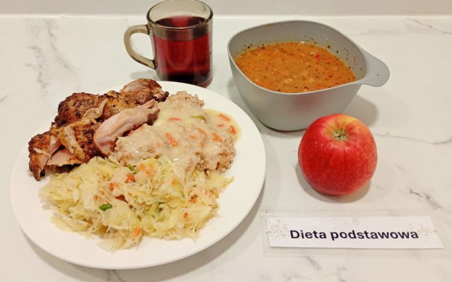 Talerz z porcją pieczonego mięsa z kurczaka, kaszą polaną jasnym sosem z warzywami oraz surówką z kapusty. Obok jabłko, miska z zupą i kubek z herbatą. Na stoliku kartka z napisem Dieta podstawowa