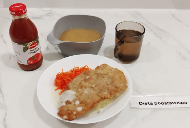 Na białym talerzu znajduje się panierowany filet z kurczaka polany jasnym sosem, porcja puree ziemniaczanego i surówka z marchewki. Obok ustawiono szklankę z ciemnym napojem, szarą miskę z zupą oraz butelkę soku pomidorowego. Na stole leży karteczka z napisem „Dieta podstawowa”
