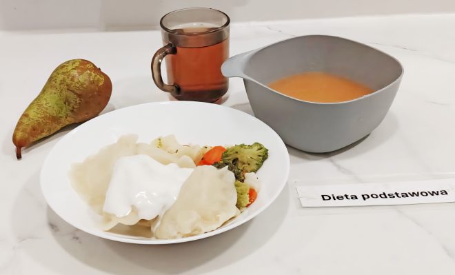 Zestaw obiadowy opisany jako „Dieta podstawowa”. Na talerzu podano pierogi polane śmietaną oraz warzywa gotowane – brokuły, marchew i kalafior. Obok znajduje się kubek z herbatą, miska z zupą pomidorową i gruszka jako dodatek. Posiłek ustawiony na białym blacie