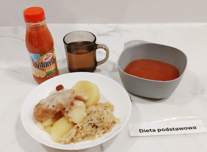 Zestaw obiadowy opisany jako „Dieta podstawowa”. Na talerzu znajdują się gotowane ziemniaki, kotlet schabowy z sosem oraz porcja duszonej kapusty. Obok stoi kubek z herbatą, miska z zupą pomidorową oraz butelka soku marchwiowo-bananowego Hortex. Wszystko ustawione na białym blacie