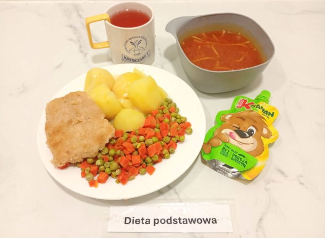 Zestaw obiadowy w diecie podstawowej. Na talerzu znajduje się panierowany filet z ryby, gotowane ziemniaki oraz mieszanka marchewki i groszku. Obok umieszczono miseczkę z zupą pomidorową z makaronem, kubek z czerwonym napojem oraz owocowy mus w saszetce. Przed zestawem widnieje etykieta z napisem „Dieta podstawowa”
