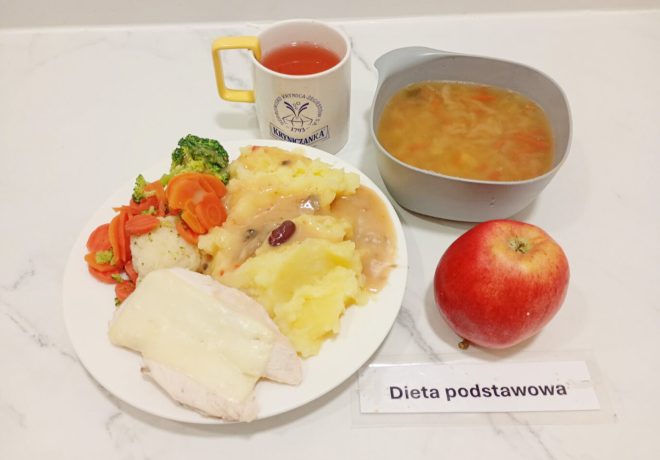 Na zdjęciu widoczny jest zestaw obiadowy w ramach diety podstawowej. Na talerzu znajduje się gotowany filet drobiowy przykryty plasterkiem sera, porcja puree ziemniaczanego z sosem pieczarkowym i warzywami strączkowymi, a obok ugotowane warzywa – marchewka, brokuł i kalafior. Do posiłku podano również zupę jarzynową w szarej miseczce, czerwone jabłko oraz kubek z herbatą owocową w żółto-białym kubku z logo „Kryniczanka”. Całość ustawiona jest na jasnym blacie, z karteczką z napisem „Dieta podstawowa”
