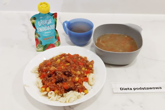 Na białym blacie ustawiono posiłek opisany jako „Dieta podstawowa”. Na talerzu znajduje się porcja makaronu świderki z sosem mięsno-warzywnym, w którym widać czerwoną fasolę, kukurydzę, paprykę i marchewkę. Obok stoi miska z zupą jarzynową, kubek z herbatą oraz saszetka musu owocowego o smaku mango i jabłka. Całość stanowi przykład obiadu w standardowej diecie szpitalnej