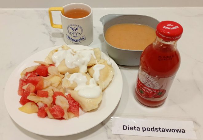 Na zdjęciu widoczny jest zestaw obiadowy oznaczony jako dieta podstawowa: talerz z pierogami polanymi sosem śmietanowym i porcją gotowanych owoców, miseczka z zupą krem, butelka soku pomidorowego oraz kubek z herbatą. Posiłek podany na białym blacie