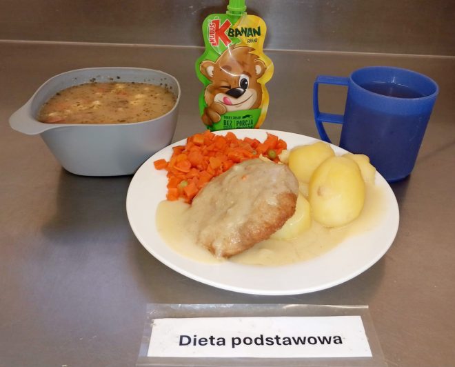 Na metalowym blacie stoi zestaw obiadowy oznaczony etykietą „Dieta podstawowa”. Na białym talerzu znajduje się porcja ziemniaków, panierowany kotlet w jasnym sosie oraz gotowana marchewka z groszkiem. Obok stoi miska z zupą — wygląda jak żurek lub zupa jarzynowa z kawałkami kiełbasy i przyprawami. W tle widoczny jest mus owocowy w saszetce marki Kubuś o smaku banana i jabłka oraz niebieski kubek z ciemnym napojem, prawdopodobnie herbatą