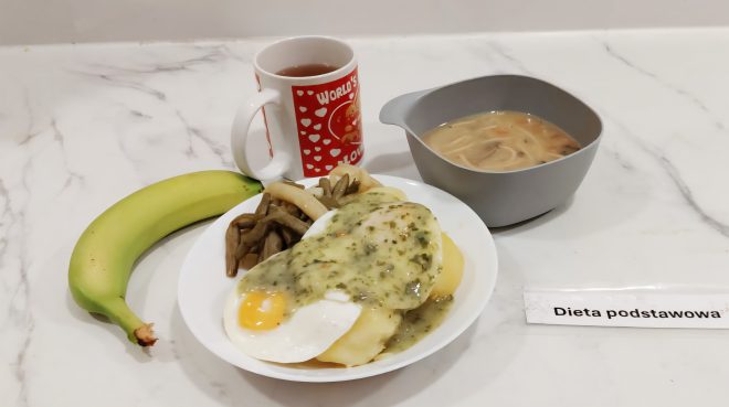 Obiad w diecie podstawowej. Na tacy zupa z makaronem, danie główne: ziemniaki z jajkiem sadzonym, zielonym sosem i fasolką szparagową, a także banan i kubek z herbatą