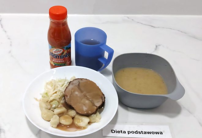 Na zdjęciu obiad w ramach diety podstawowej: porcja pieczeni z sosem, kopytka i surówka z białej kapusty. Obok szara miseczka z zupą jarzynową oraz napój w niebieskim kubku. Z tyłu butelka soku marchwiowo-brzoskwiniowego „Vitaminka”. Na stole kartka z napisem „Dieta podstawowa”