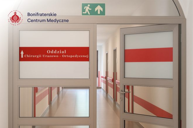 Przeszklone drzwi prowadzące na Oddział Chirurgii Urazowo-Ortopedycznej w Bonifraterskim Centrum Medycznym. Na szybie widoczny czerwony pasek z napisem „Oddział Chirurgii Urazowo-Ortopedycznej”, a nad drzwiami znak ewakuacyjny z zieloną strzałką. Za drzwiami jasny, schludny korytarz z czerwonymi poręczami i kilkoma wejściami do sal