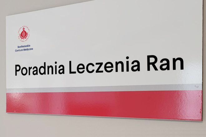 Tabliczka informacyjna z napisem „Poradnia Leczenia Ran” umieszczona na ścianie. W lewym górnym rogu widoczne jest logo Bonifraterskiego Centrum Medycznego. Tabliczka utrzymana jest w biało-czerwonej kolorystyce z szarym paskiem oddzielającym część tekstową od dolnej czerwonej belki