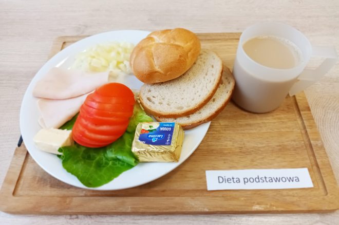 Śniadanie: dwa plasterki szynki drobiowej, pomidor, sałata, masło i serek topiony. Obok bułka, dwie kromki chleba pszennego oraz kubek kawy z mlekiem. Na stoliku kartka z napisem Dieta podstawowa