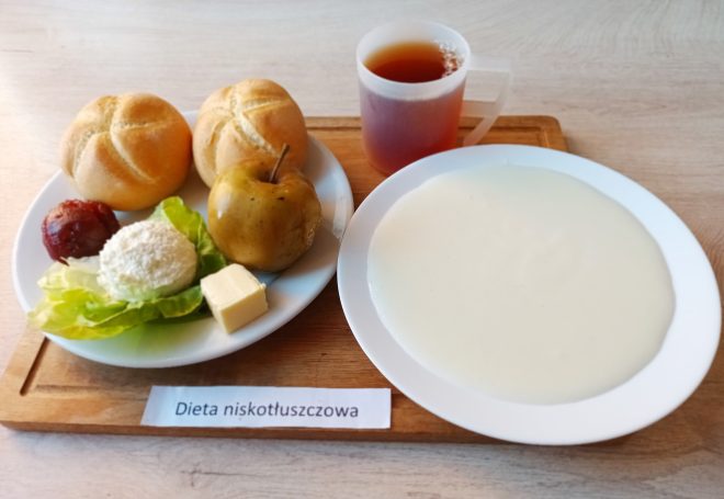 Śniadanie: dwa kajzerki, jabłko, plaster sera żółtego, twarożek i plaster kiełbasy na sałacie. Obok talerz z mleczną zupą kremową oraz kubek herbaty. Na stoliku kartka z napisem Dieta niskotłuszczowa