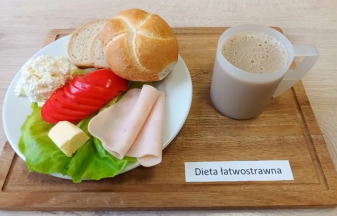 Zestaw śniadaniowy w ramach diety łatwostrawnej, podany na drewnianej tacy. Na talerzu znajdują się: bułka kajzerka, dwie kromki pieczywa pszennego, plasterki pomidora ułożone na liściu sałaty, dwa plastry wędliny drobiowej, kostka masła oraz porcja białego twarogu. Obok talerza stoi kubek z jasnym napojem mlecznym lub kawą z mlekiem