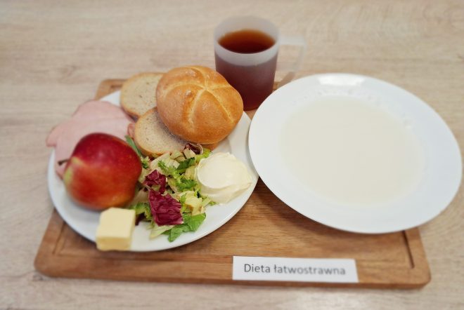 Na tacy z napisem „Dieta łatwostrawna” znajduje się zestaw śniadaniowy: na białym talerzu położono świeżą bułkę, dwa plasterki pieczywa pszennego, kilka plasterków chudej wędliny drobiowej, porcję sałatki z liści sałaty i radicchio z kleksem jogurtu naturalnego, kostkę masła oraz czerwone jabłko, obok talerza stoi filiżanka z herbatą oraz miska z jasną zupą mleczną. Całość znajduje się na drewnianej tacy