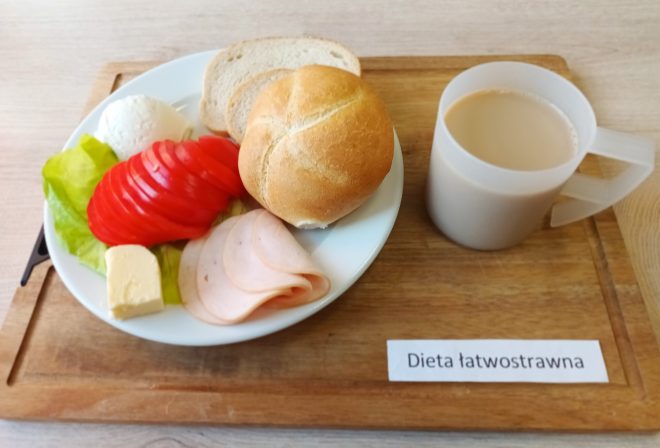 Na tacy z napisem „Dieta łatwostrawna” znajduje się śniadanie: na talerzu widoczna jest bułka kajzerka, kromka chleba pszennego, kilka plasterków chudej wędliny drobiowej, kawałek masła, liść sałaty, pokrojony pomidor i kulka białego twarożku. Obok ustawiono kubek z jasną kawą z mlekiem