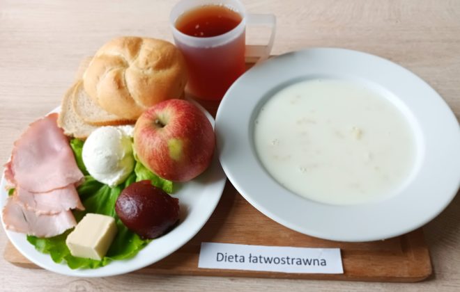 Taca z posiłkiem oznaczona etykietą „Dieta łatwostrawna”. Na talerzu widoczna jest bułka, kromka jasnego pieczywa, plasterki chudej szynki, twarożek, kawałek masła, galaretka owocowa oraz jabłko ułożone na liściu sałaty. Obok znajduje się talerz z mleczną zupą z drobną kaszką i kubek z herbatą