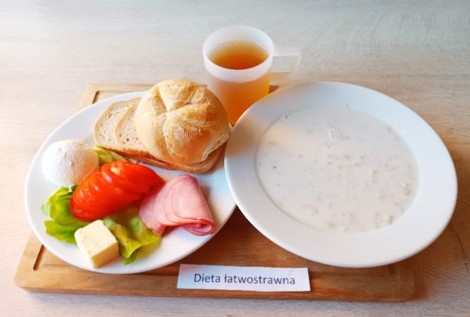 Zestaw śniadaniowy w diecie łatwostrawnej. Na talerzu bułka i kromki chleba, plasterki pomidora, szynka, kostka masła, kulka białego sera i liść sałaty. Obok talerza miska z mleczną zupą oraz kubek z herbatą. Na tabliczce widoczny napis „Dieta łatwostrawna”