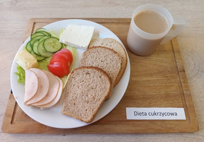 Taca z posiłkiem oznaczonym jako „Dieta cukrzycowa”. Na talerzu trzy kromki chleba pełnoziarnistego, kilka plasterków wędliny drobiowej, plaster białego sera, masło, plasterki ogórka i pomidora ułożone na liściu sałaty. Obok talerza plastikowy kubek z kawą z mlekiem