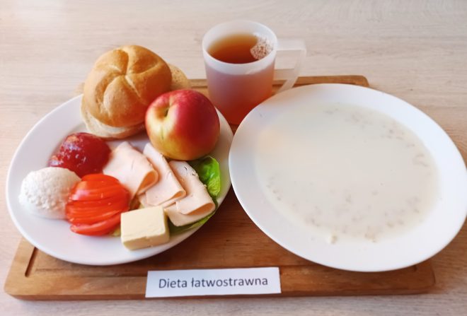 Śniadanie na białym talerzu: bułka kajzerka, jabłko, plasterki szynki drobiowej, masło, pomidor, biały ser i porcja dżemu. Obok filiżanka z herbatą oraz talerz z mleczną zupą. Całość na drewnianej tacy z etykietą „Dieta łatwostrawna”