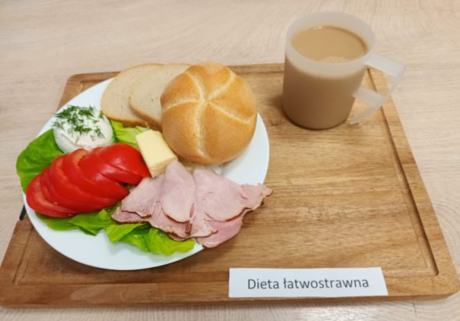 Śniadanie w diecie łatwostrawnej. Na drewnianej tacy ustawiono talerz z bułką kajzerką, kromką chleba pszennego, plastrem masła, kilkoma plastrami szynki, pomidorem w plasterkach, sałatą i kulką białego twarożku z koperkiem. Obok przezroczysty kubek z kawą z mlekiem