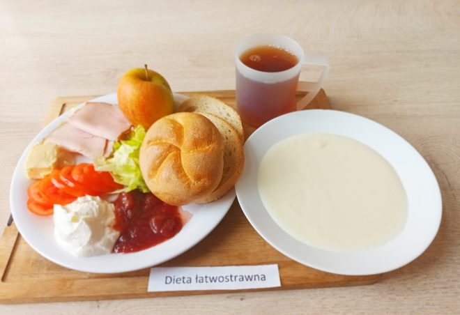 Na tacy podano śniadanie w diecie łatwostrawnej: kromki pieczywa, bułkę kajzerkę, plasterki pomidora, sałatę, masło, wędlinę drobiową, twarożek, porcję dżemu, jabłko oraz kubek z herbatą. Obok na talerzu znajduje się zupa mleczna lub kremowa kasza manna. Pod etykietą napis: „Dieta łatwostrawna”