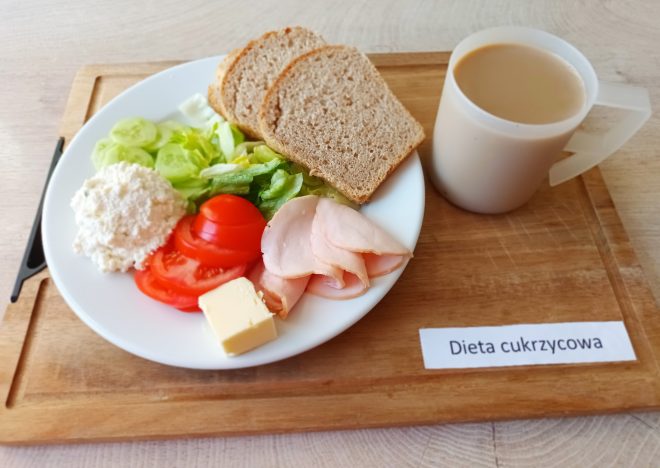 Na drewnianej tacy znajduje się śniadanie oznaczone etykietą „Dieta cukrzycowa”. Na talerzu podano trzy kromki pieczywa pełnoziarnistego, plasterki drobiowej wędliny, porcję twarogu, kilka plastrów pomidora i ogórka oraz listki sałaty. Obok leży mała kostka masła. Do zestawu dołączony jest kubek z kawą z mlekiem. Posiłek wygląda lekko i zbilansowanie, odpowiedni dla osób z cukrzycą