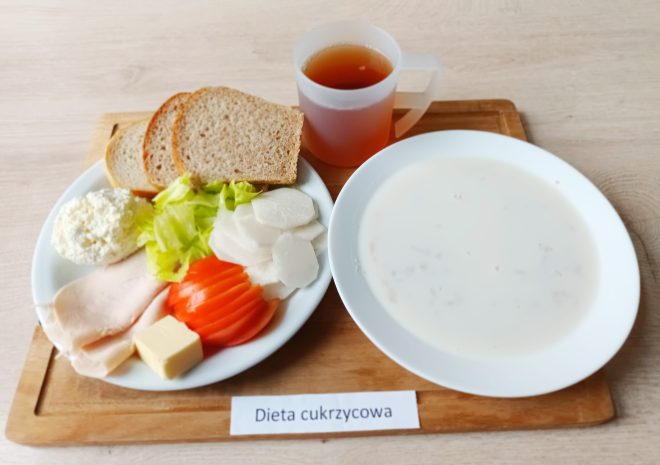Na tacy ustawionej na drewnianej desce znajduje się posiłek opisany jako „Dieta cukrzycowa”. Na białym talerzu widać trzy kromki chleba razowego, plasterki pomidora, sałatę, plasterki białego warzywa (najprawdopodobniej rzodkiew lub kalarepa), plaster chudej wędliny drobiowej, porcję białego twarożku i kostkę masła. Obok stoi miska z mleczną zupą oraz kubek z ciemną herbatą