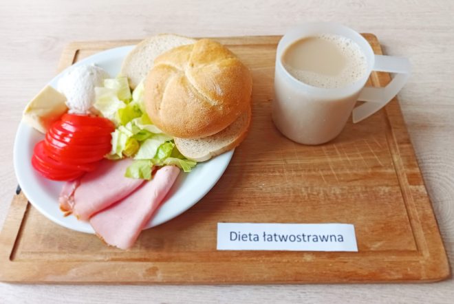 Na drewnianej tacy podano śniadanie w ramach diety łatwostrawnej: bułka kajzerka z masłem, plasterkami szynki, sałatą, pomidorem i twarożkiem. Obok kubek z kawą z mlekiem. Danie ułożone estetycznie na białym talerzu