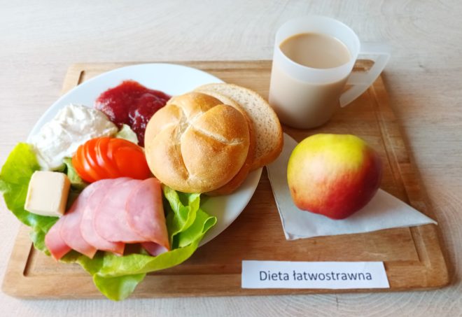 Śniadanie w diecie łatwostrawnej. Na talerzu bułka i kromki chleba z szynką, twarożkiem, pomidorem i sałatą, porcja dżemu i kostka masła. Obok kubek z kawą z mlekiem i jabłko